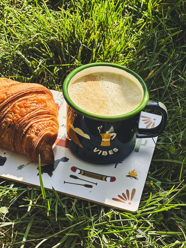 grüne Emaille Tasse mit aufdruck auf einem Schneidebrett mit einem Croissant und Wiese
